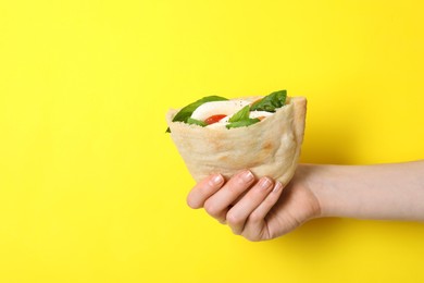 Woman holding delicious pita sandwich with mozzarella, tomatoes and basil on yellow background, closeup