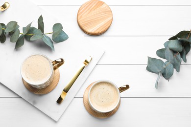 Mugs of coffee with stylish cup coasters and eucalyptus branches on white wooden table, flat lay