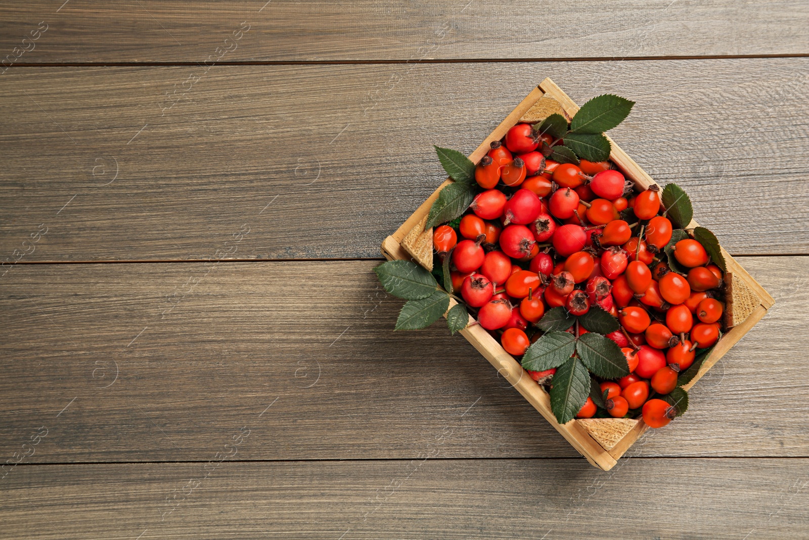 Photo of Ripe rose hip berries with green leaves in crate on wooden table, top view. Space for text