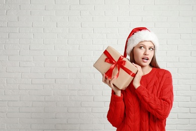 Emotional young woman in Santa hat with Christmas gift near white brick wall. Space for text