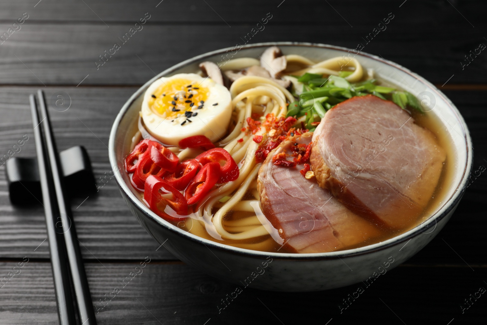 Photo of Delicious ramen in bowl and chopsticks on black wooden table, closeup. Noodle soup