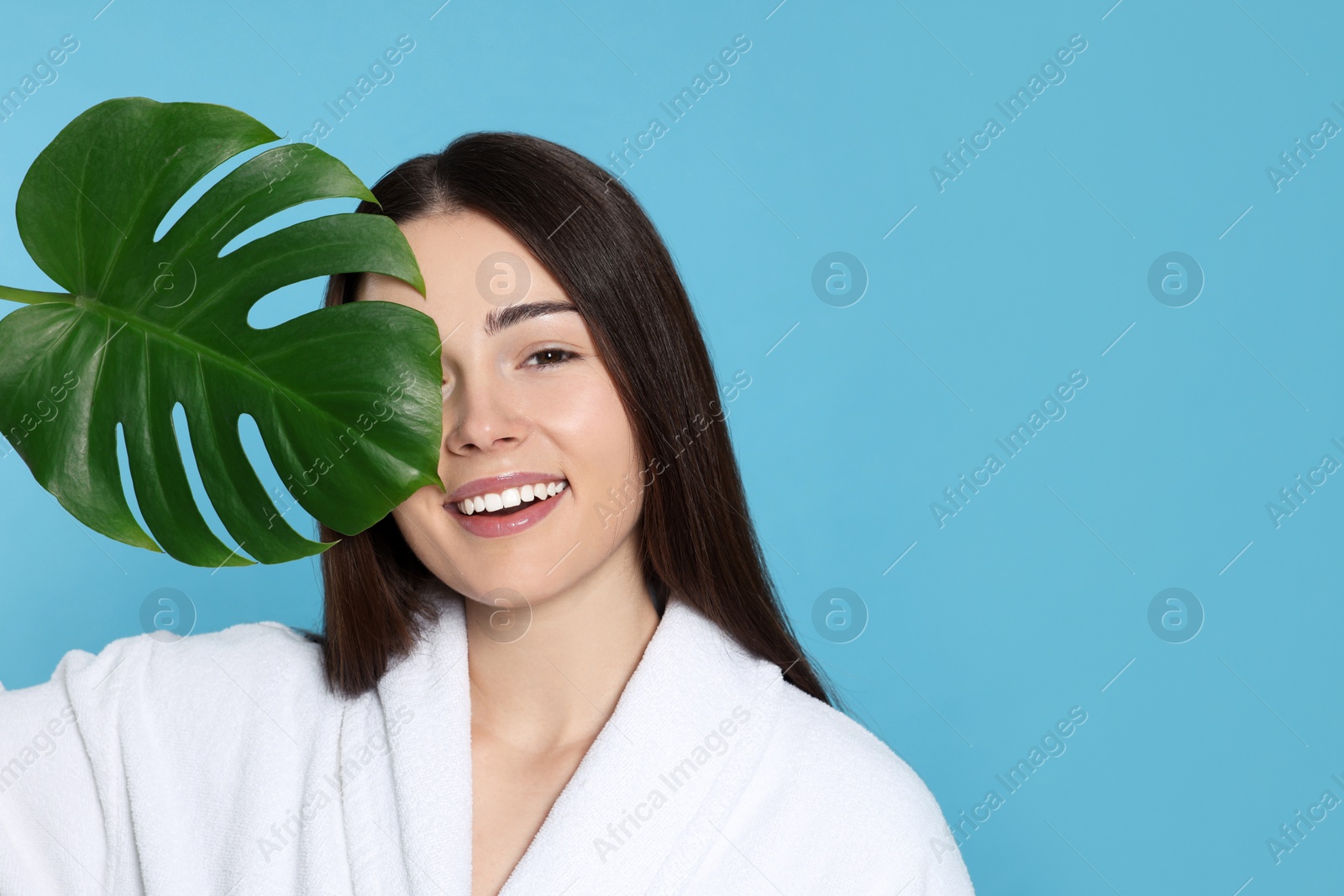 Photo of Woman in bathrobe holding leaf of monstera on light blue background, space for text. Spa treatment