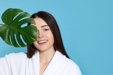 Woman in bathrobe holding leaf of monstera on light blue background, space for text. Spa treatment