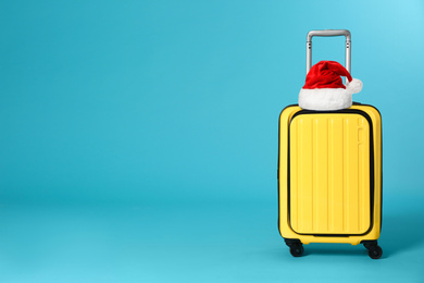 Image of Stylish suitcase with Santa Claus hat on blue background, space for text. Christmas vacation  