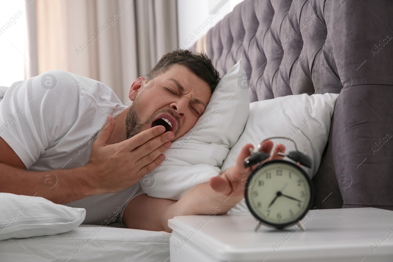 Photo of Sleepy man turning off alarm clock at home in morning