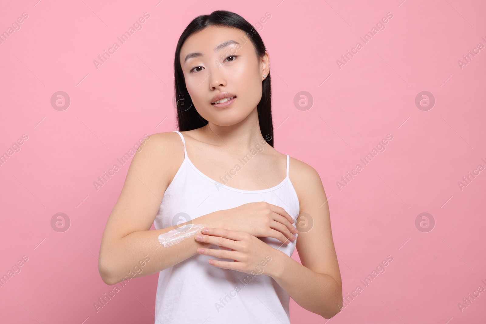 Photo of Beautiful young Asian woman applying body cream onto arm on pink background