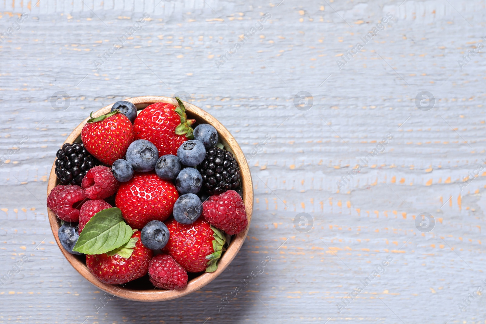 Photo of Different fresh ripe berries in bowl on light grey wooden table, top view. Space for text