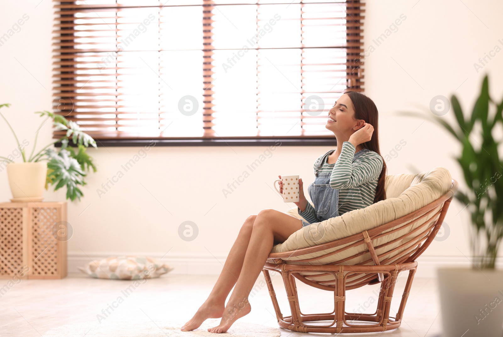 Photo of Young woman relaxing in papasan chair near window with blinds at home. Space for text