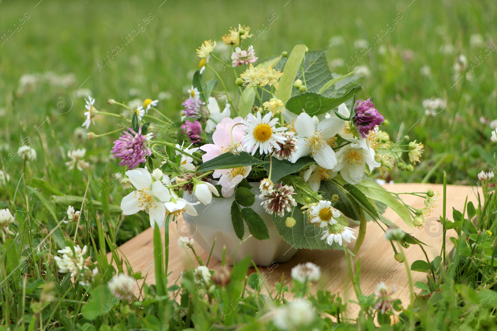 Photo of Ceramic mortar with different wildflowers and herbs on wooden board in meadow