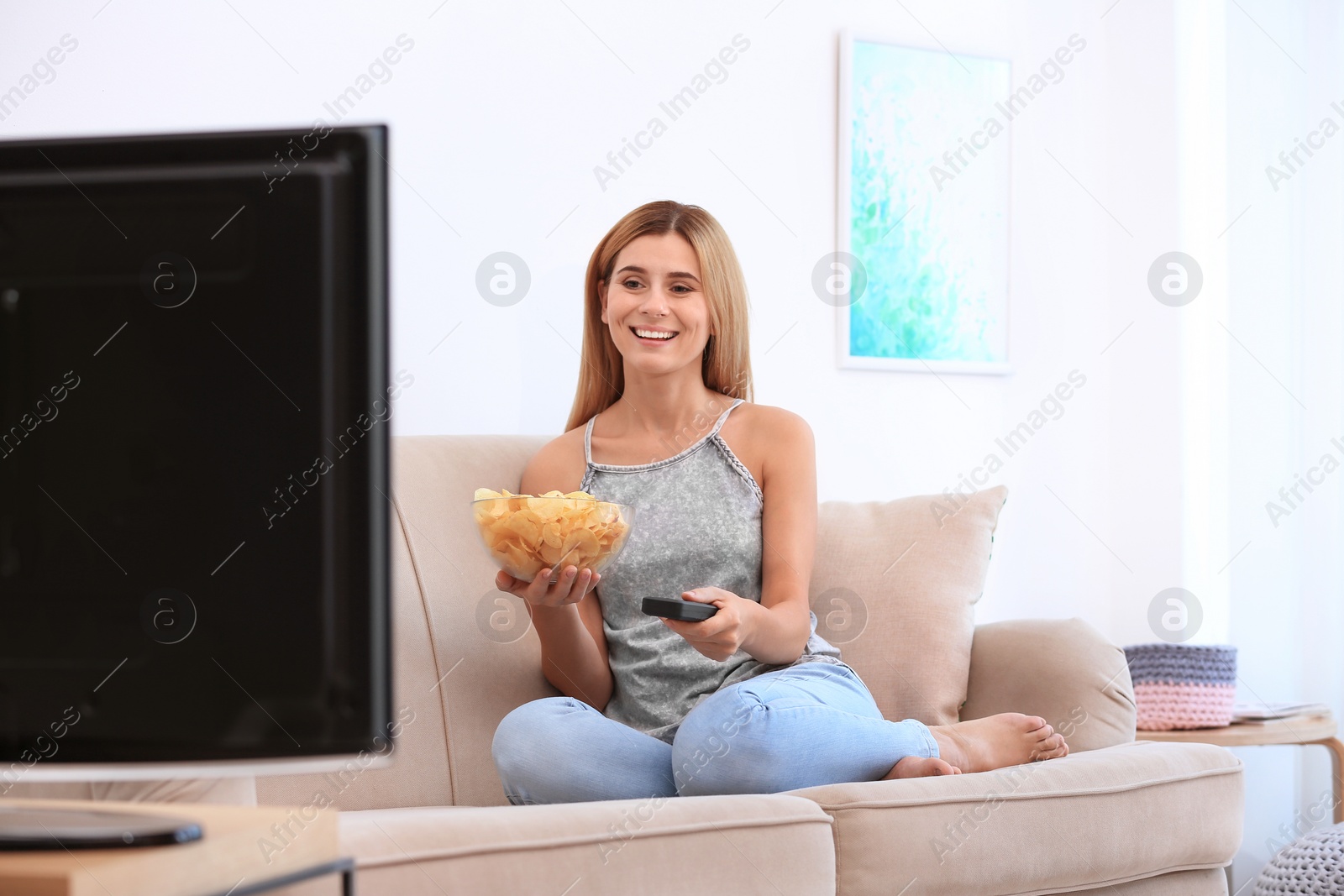 Photo of Woman with bowl of potato chips watching TV on sofa in living room