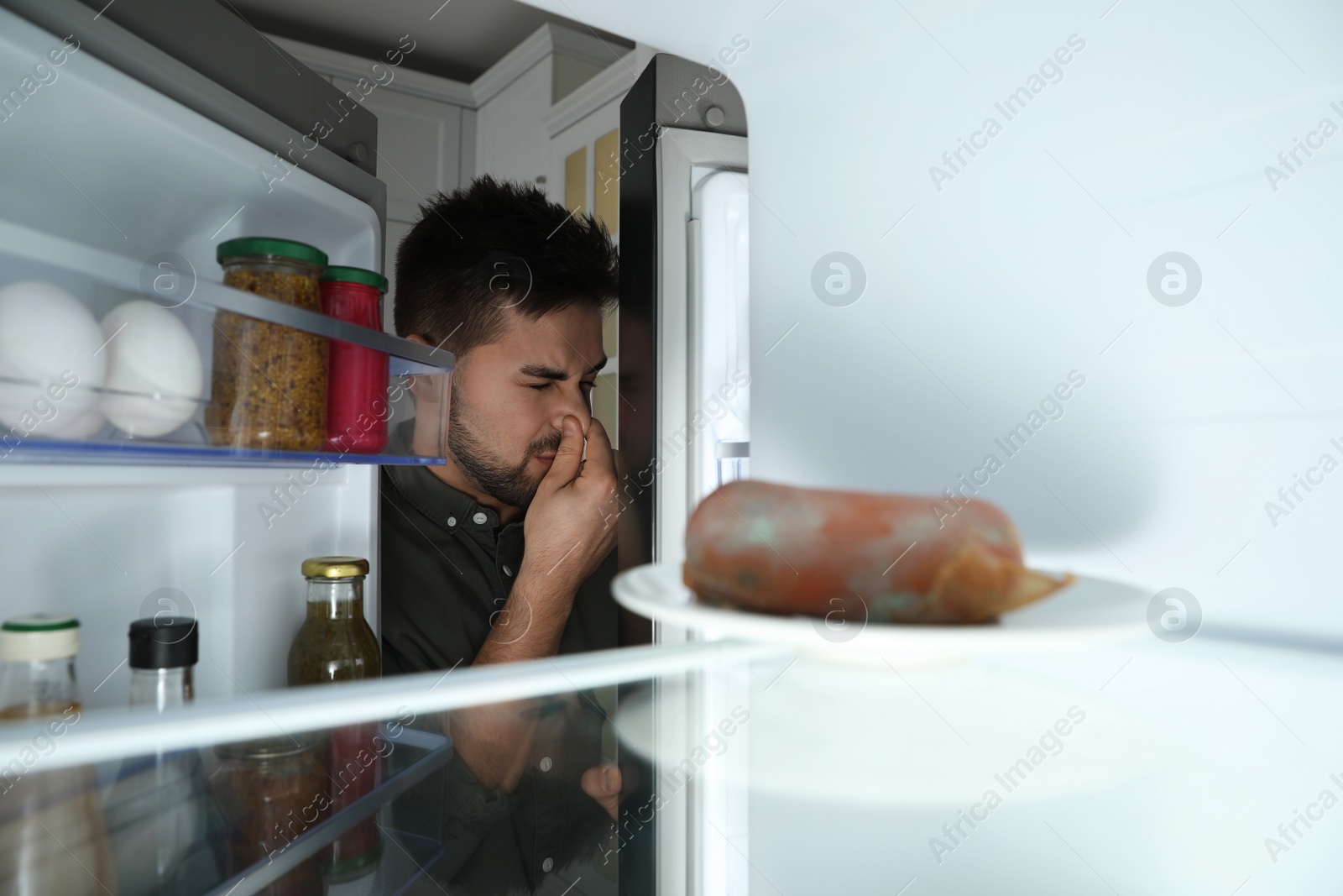 Photo of Young man feeling bad smell of spoiled sausage in refrigerator, view from inside