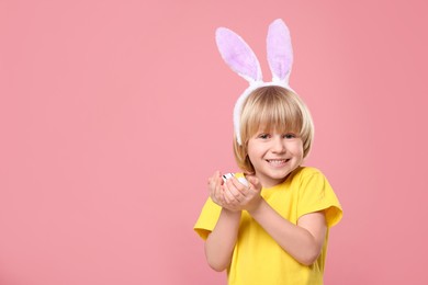 Happy boy in bunny ears headband holding painted Easter eggs on pink background. Space for text