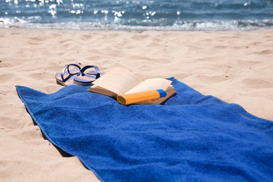 Photo of Soft blue beach towel, flip flops, sunblock and book on sandy seashore