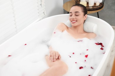 Photo of Happy woman taking bath in tub with foam and rose petals indoors