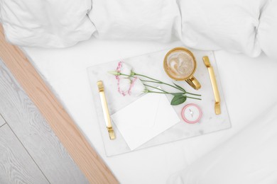 Photo of Tray with cup of coffee, flowers and beautiful engagement ring in box on white bed, top view