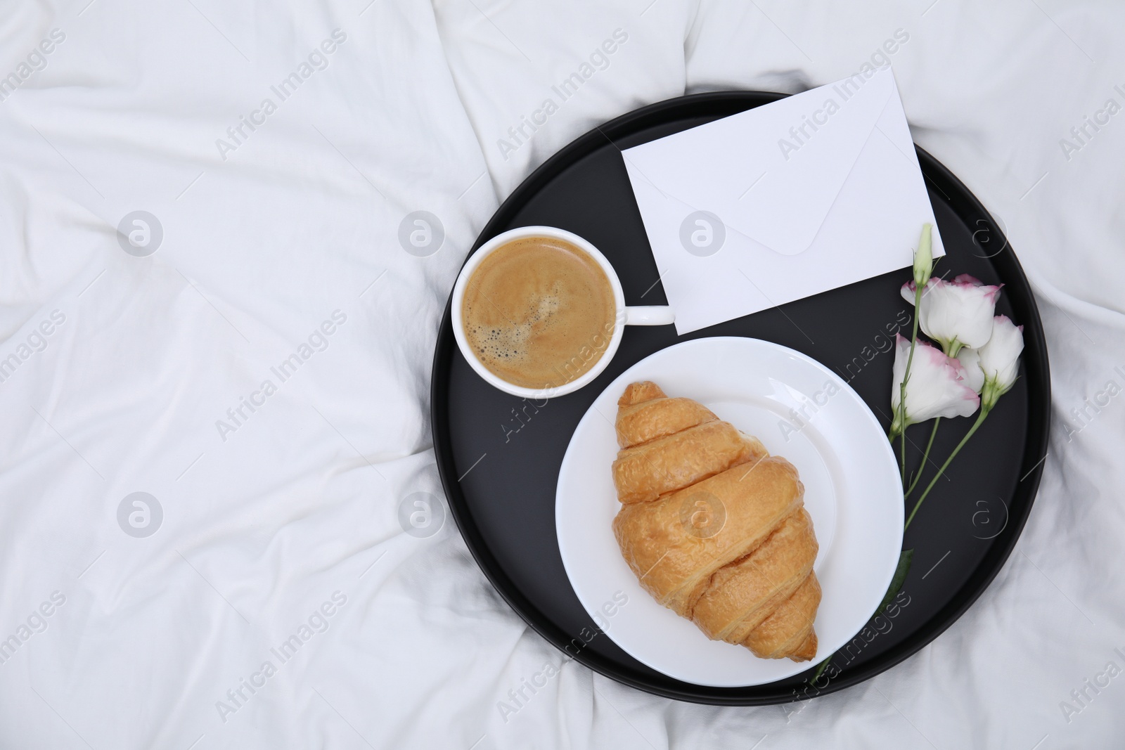 Photo of Tray with tasty croissant, cup of coffee and flowers on white bed, top view. Space for text