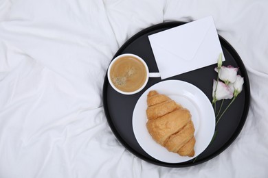 Photo of Tray with tasty croissant, cup of coffee and flowers on white bed, top view. Space for text