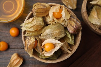 Ripe physalis fruits with calyxes in bowl on wooden table, flat lay