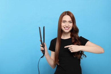Photo of Beautiful woman pointing at hair iron on light blue background. Space for text