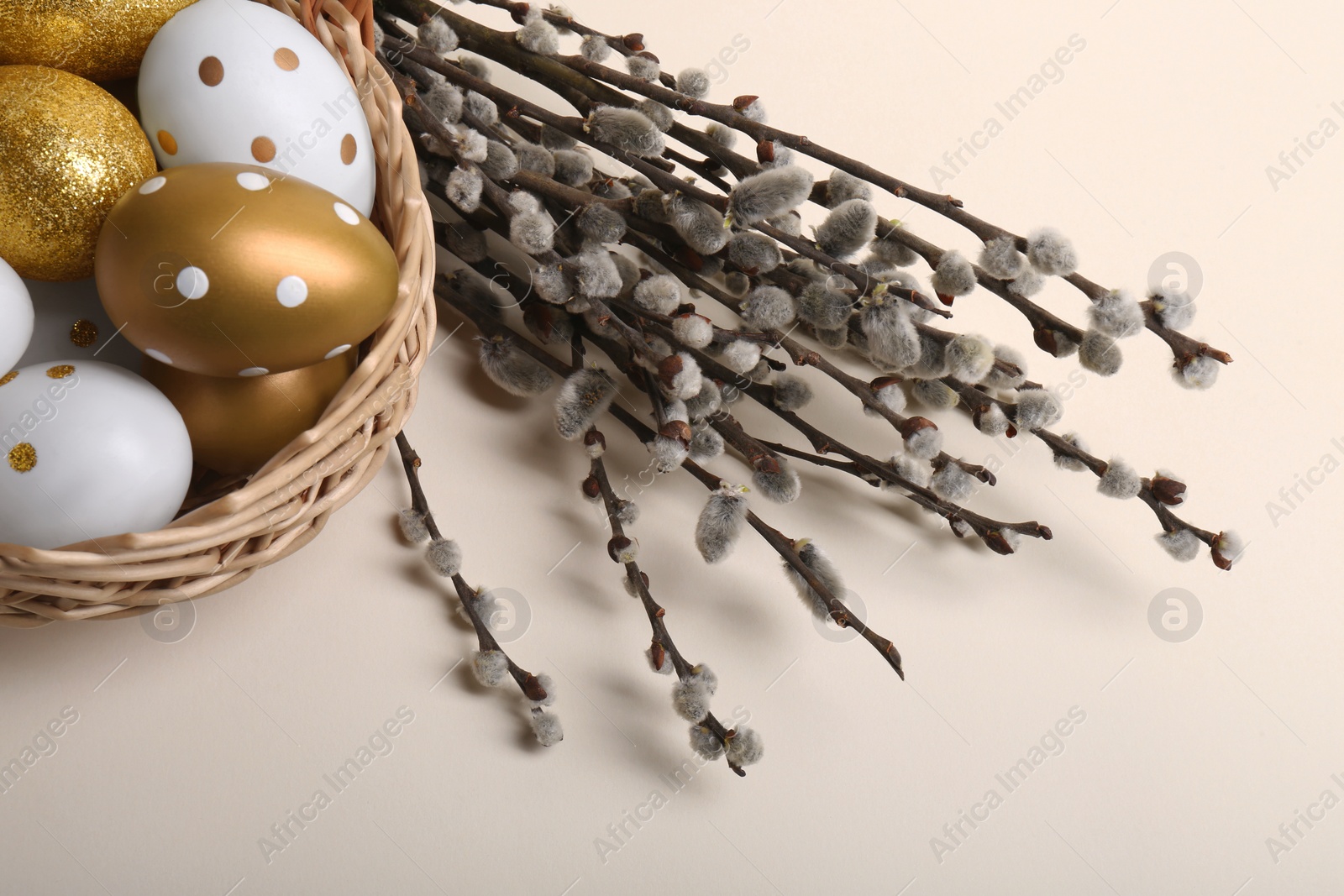 Photo of Wicker basket with decorated Easter eggs and willow branches on beige background, closeup