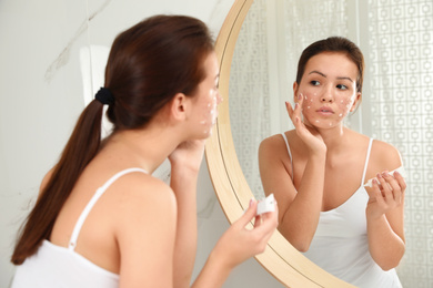 Photo of Teen girl with acne problem applying cream near mirror in bathroom