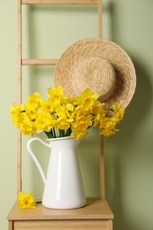 Jug with beautiful daffodils and wicker hat on wooden chair near light green wall