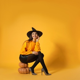 Photo of Beautiful woman wearing witch costume for Halloween party sitting on pumpkins against yellow background, space for text