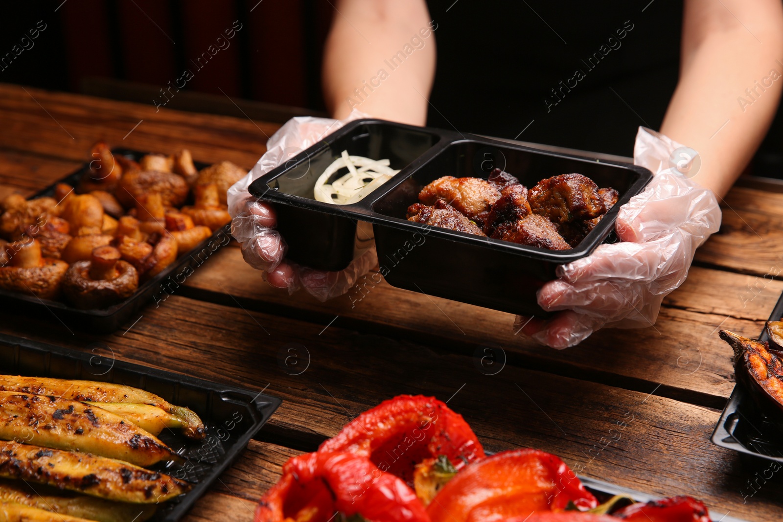 Photo of Waiter with plastic container of tasty shish kebab at wooden table, closeup. Food delivery service