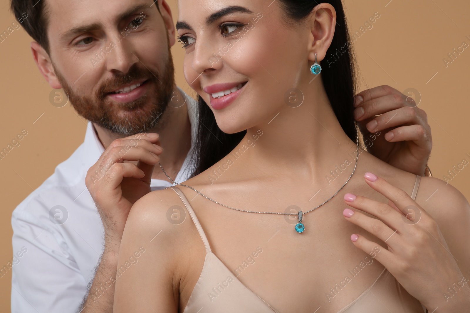 Photo of Man putting elegant necklace on beautiful woman against beige background, closeup