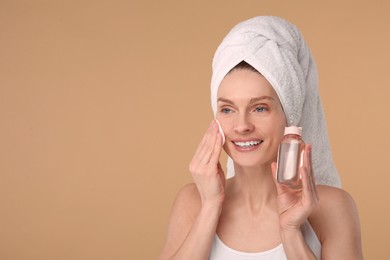 Beautiful woman in terry towel removing makeup with cotton pad on beige background, space for text