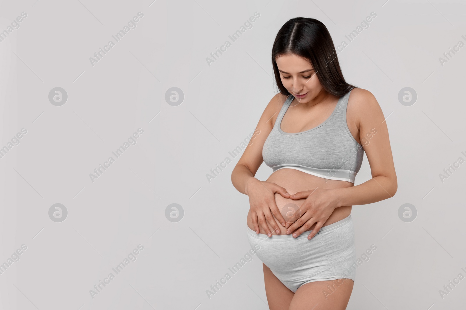 Photo of Beautiful pregnant woman in comfortable maternity underwear making heart with hands on her belly against grey background, space for text
