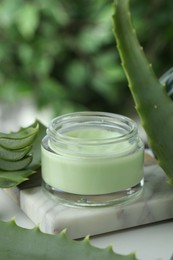 Jar with cream and cut aloe leaves on white table against blurred green background, closeup