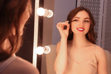 Beautiful young woman in elegant dress applying mascara near mirror indoors