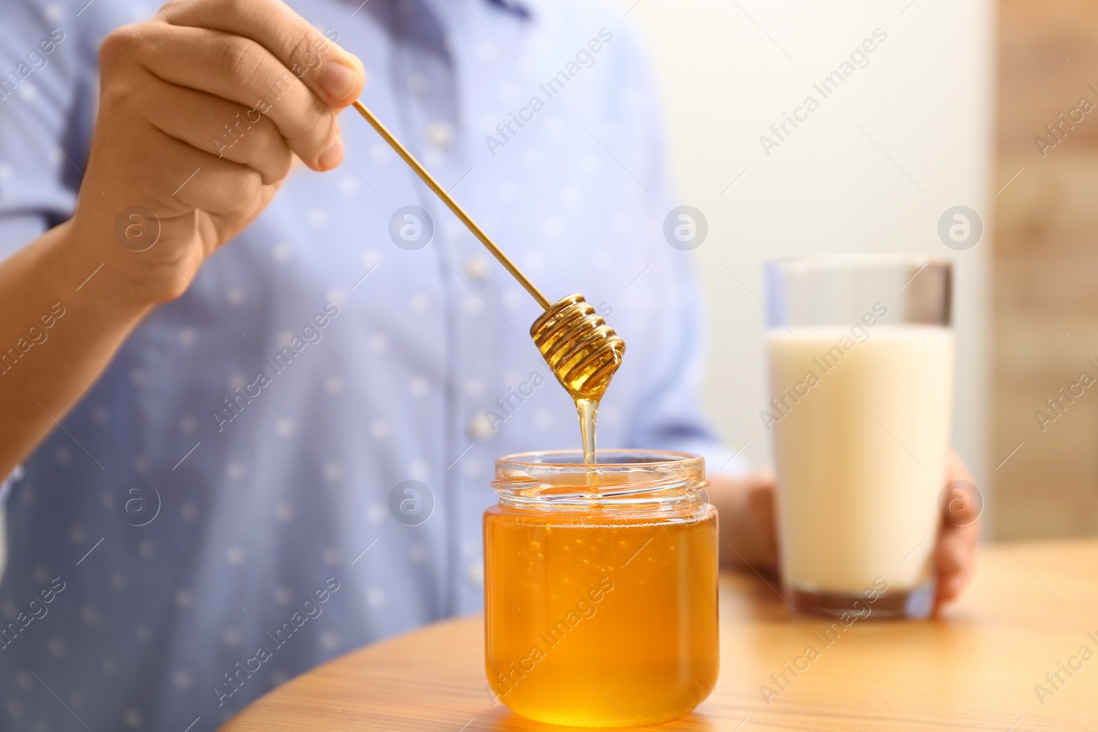 Photo of Woman holding honey dipper over jar at wooden table, closeup