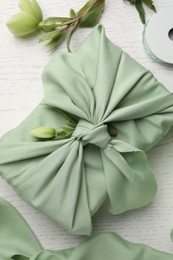 Photo of Furoshiki technique. Gift packed in green fabric and decorated with hellebore flower on white wooden table, flat lay