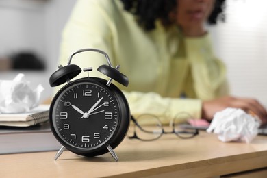 Photo of Deadline concept. Black alarm clock near woman working at wooden desk indoors, selective focus. Space for text