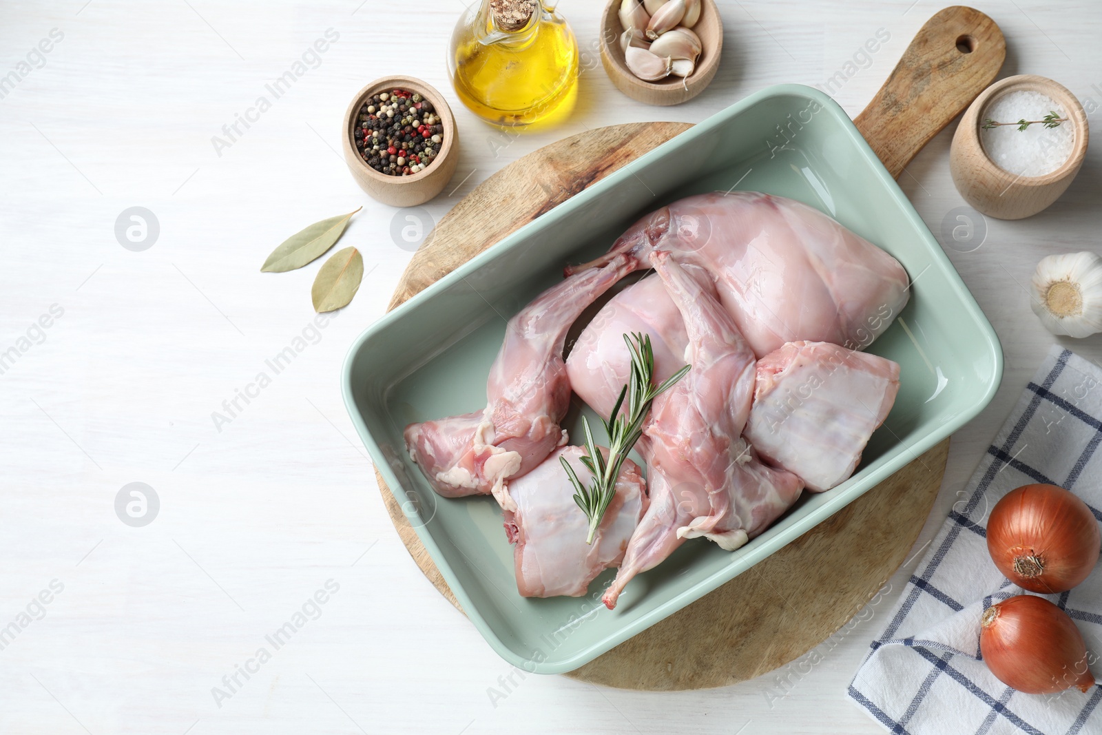 Photo of Raw rabbit meat in baking dish and products on white wooden table, flat lay. Space for text