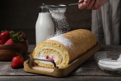 Photo of Woman pouring powdered sugar onto delicious sponge cake roll with strawberries and cream at wooden table, closeup