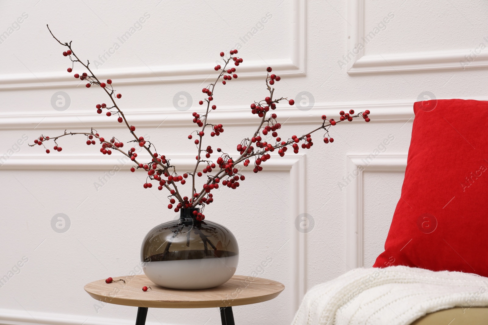 Photo of Hawthorn branches with red berries in vase on table near white wall indoors