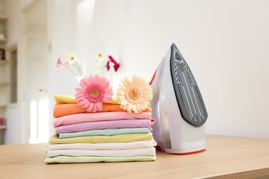 Photo of Stack of clean clothes, modern iron and flowers on wooden table