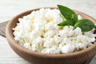 Photo of Delicious cottage cheese with basil in wooden bowl on white table, closeup