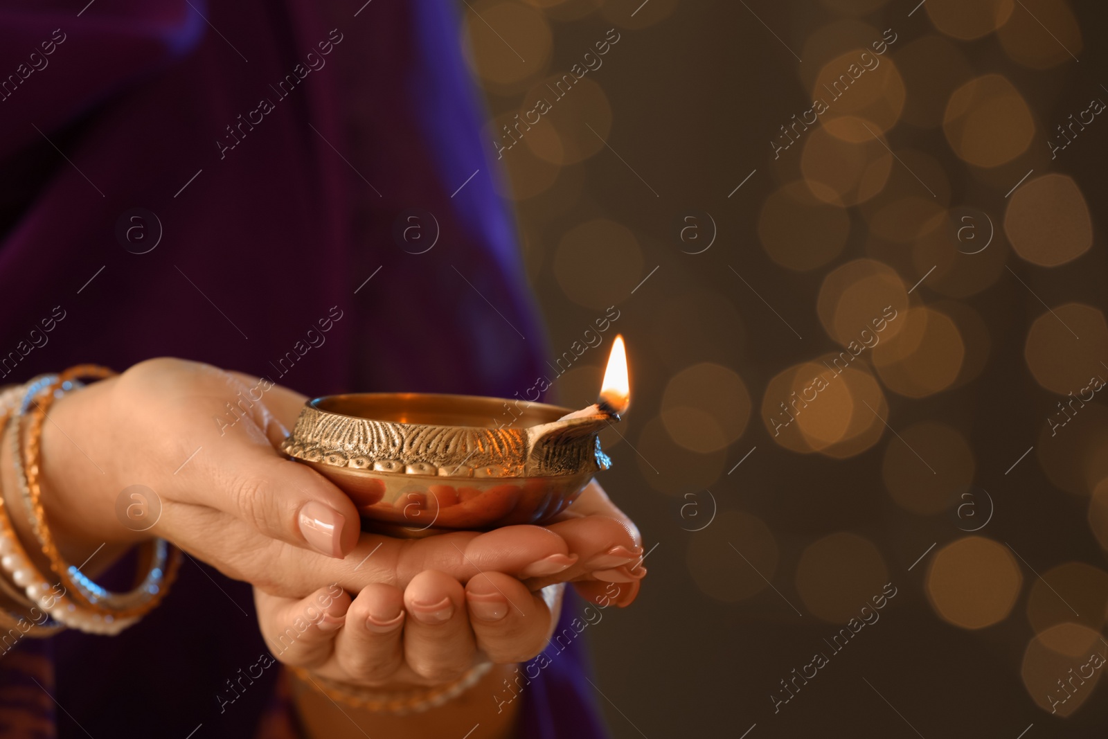 Photo of Woman holding lit diya lamp in hands against blurred lights, closeup. Diwali celebration