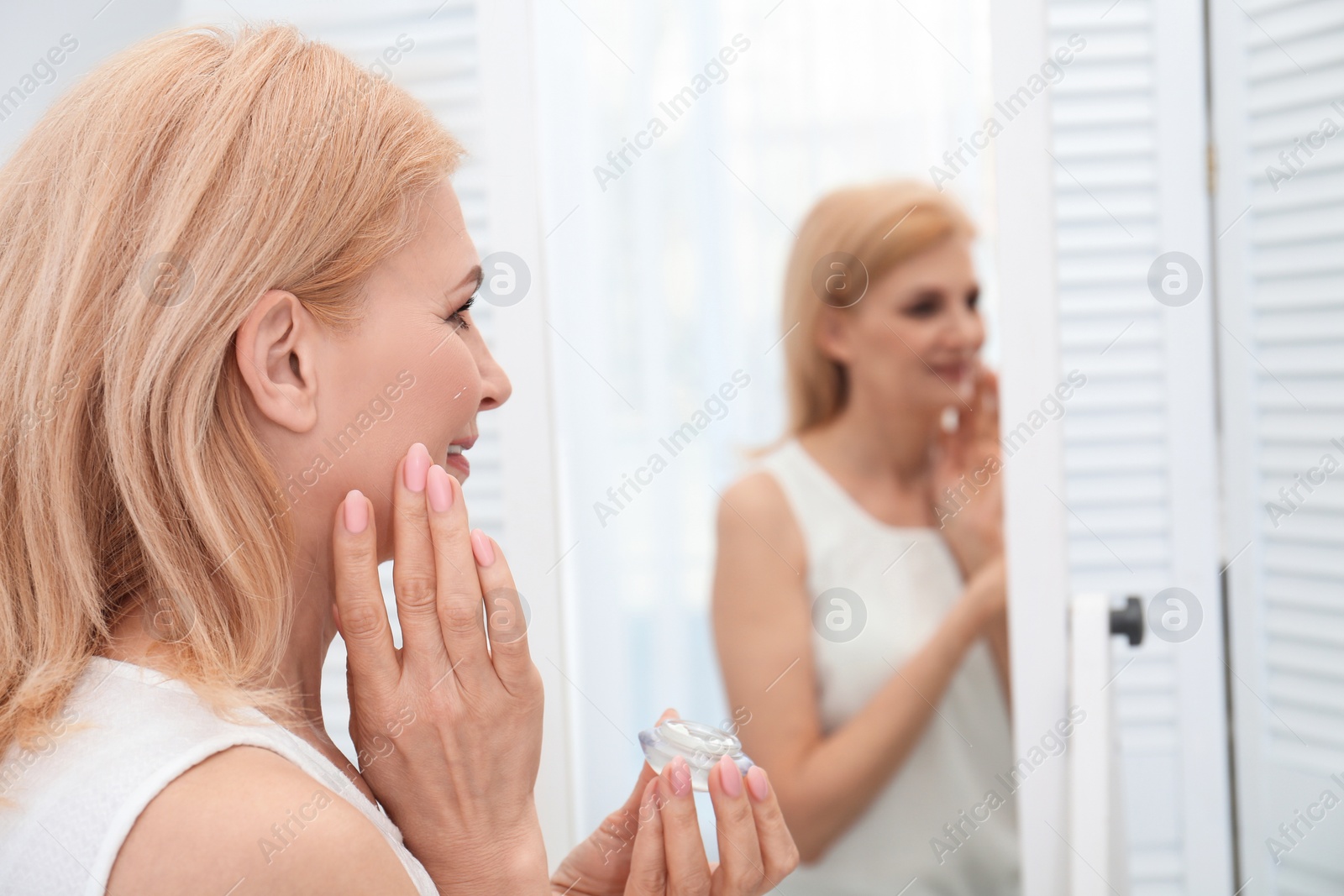 Photo of Mature woman applying face cream near mirror indoors. Space for text
