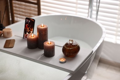 Photo of White wooden tray with burning candles, smartphone and comb on bathtub in bathroom