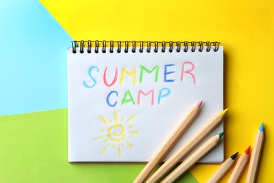 Notebook with written text SUMMER CAMP and different pencils on color background, top view