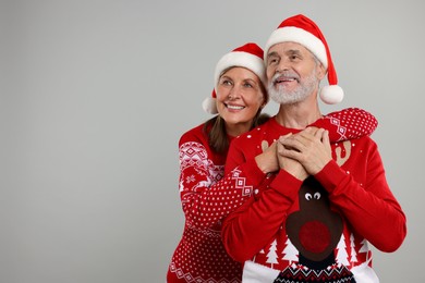 Happy senior couple in Christmas sweaters and Santa hats on grey background. Space for text