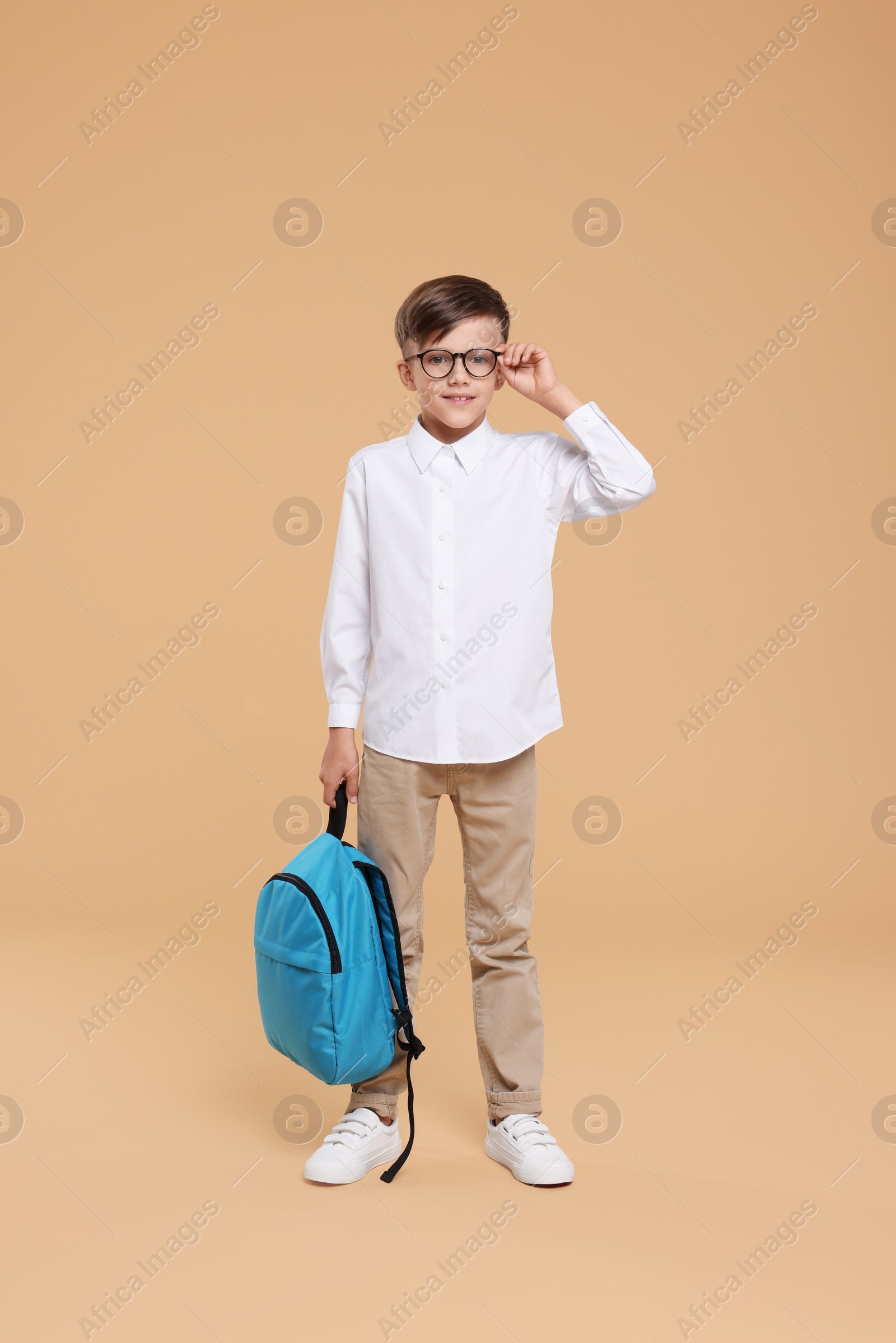 Photo of Cute schoolboy in glasses holding backpack on beige background