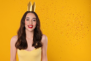 Photo of Beautiful young woman with princess crown under falling confetti on yellow background, space for text