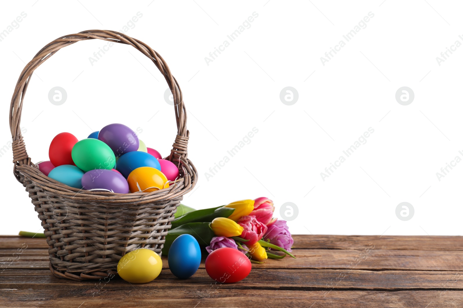 Photo of Colorful Easter eggs in wicker basket and tulips on wooden table against white background