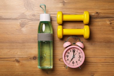 Photo of Alarm clock, dumbbells and bottle of water on wooden table, flat lay. Morning exercise